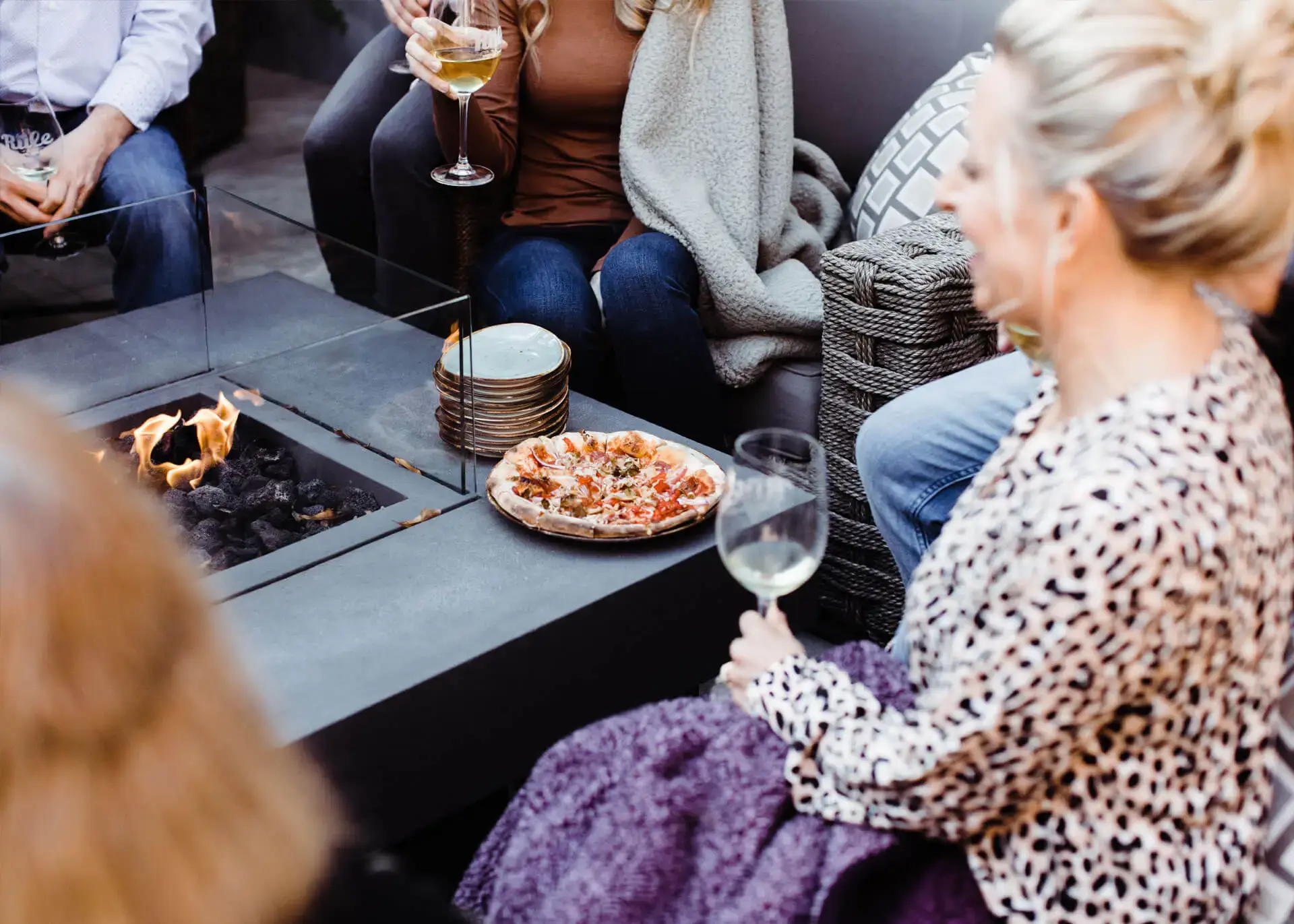 happy people gathered around firepit table patio with champagne glasses and toasty pizza having a good time